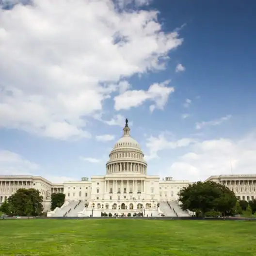 View of the United States Capitol building on a clear day, with the dome centered and surrounded by expansive green lawns.