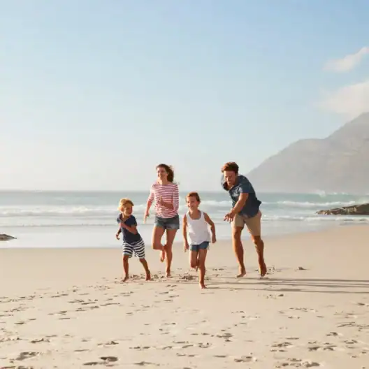 A family of four, two adults and two children, walks along a sandy beach with ocean waves in the background and mountains in the distance.