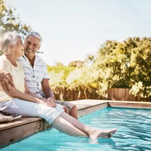 An elderly couple sits by a pool, smiling and dipping their feet in the water on a sunny day.