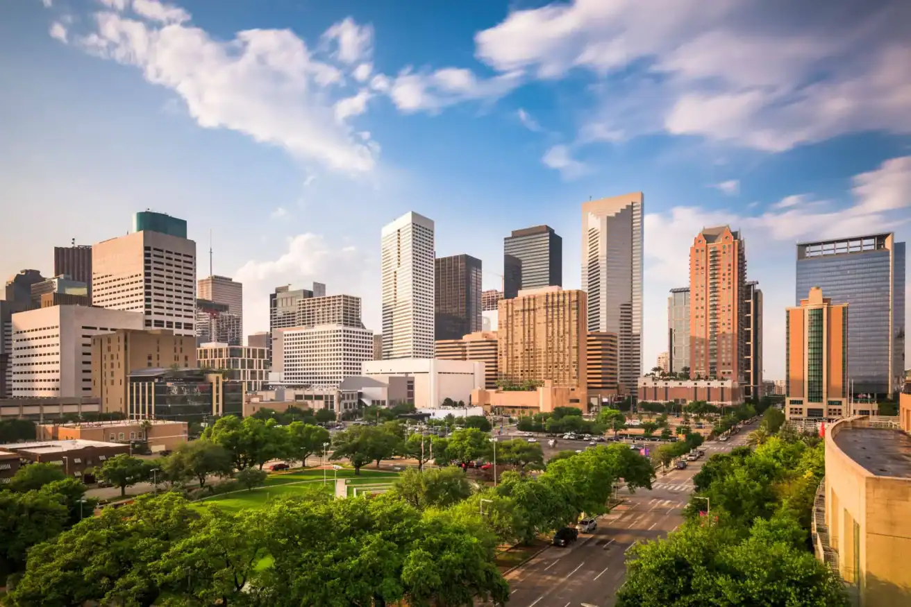 Skyline view of a city with modern skyscrapers under a partly cloudy sky, surrounded by green trees and a busy street in the foreground.