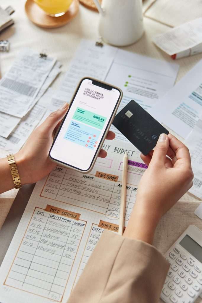Unknown woman holds smartphone and credi card enters Internet banking system pays bills on web surrounded by papers uses electronic banking services enters information. Instant epay concept.